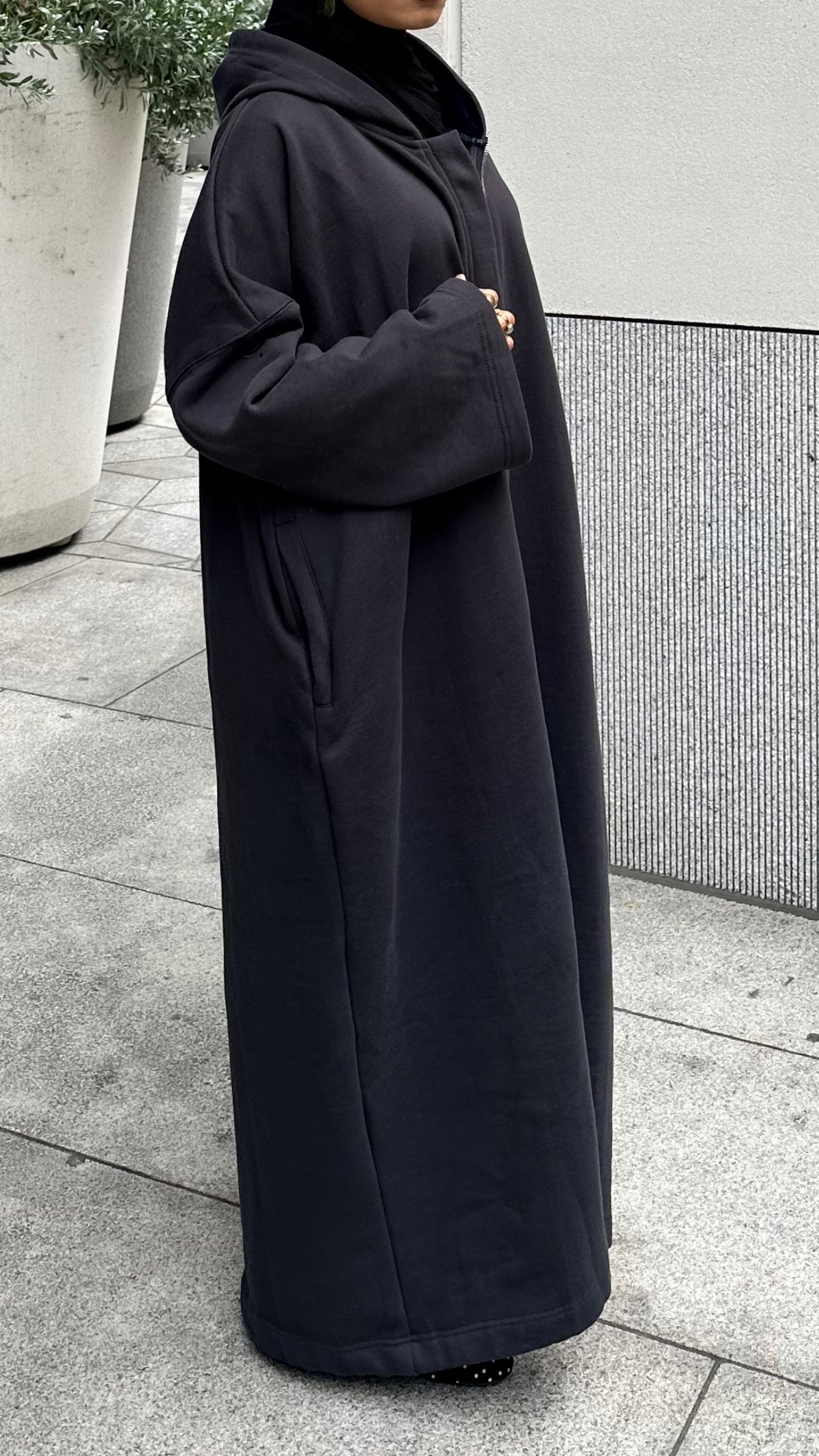 A person wearing a long black hooded coat or abaya standing outdoors on a paved walkway next to a modern wall and planters. Winter abaya perfect for cold winters