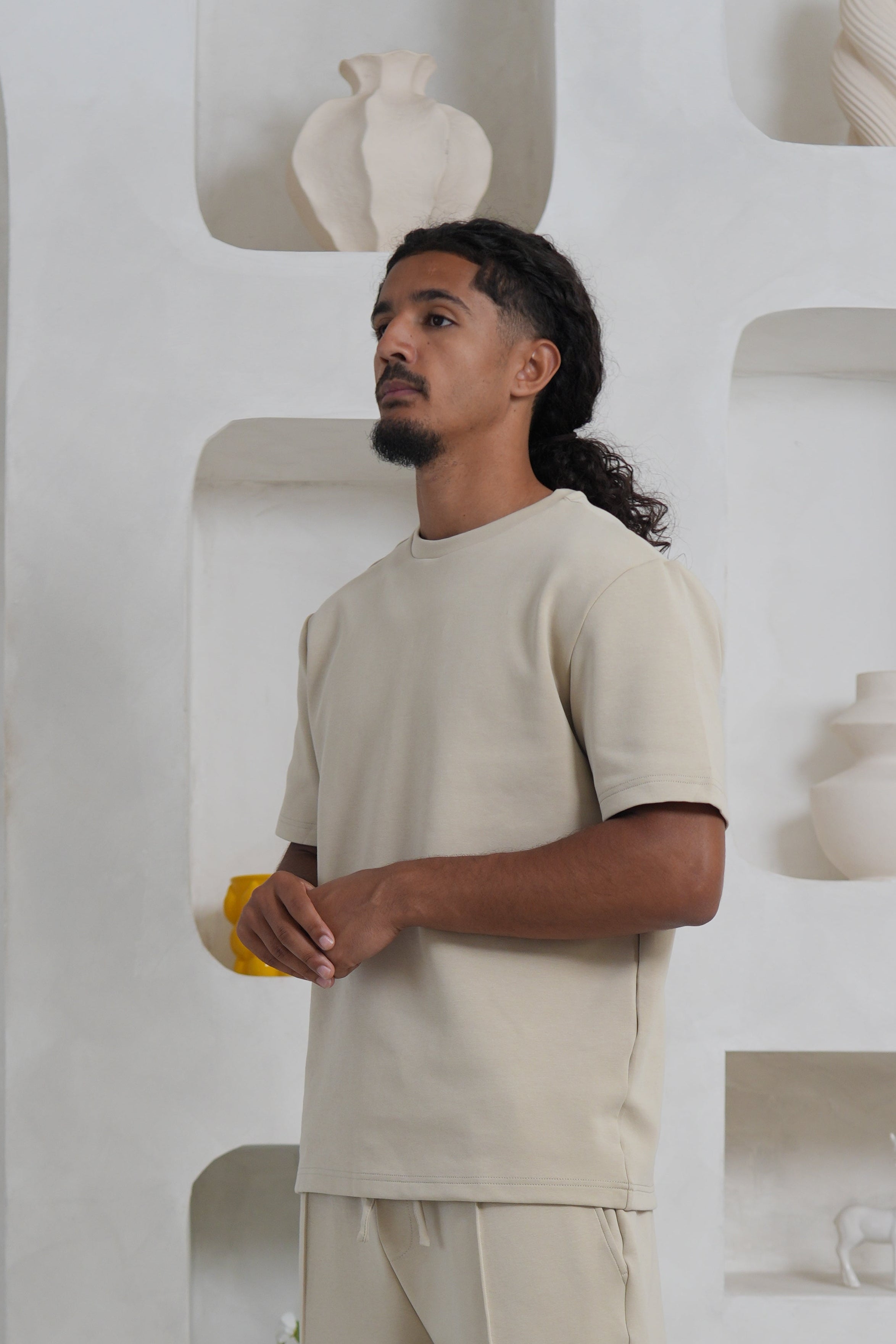 Person in a beige outfit standing in a minimalistic room with white walls and floor.