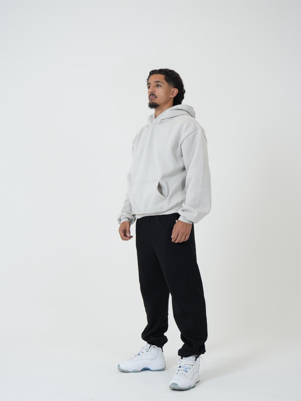 A man wearing a light gray pullover hoodie, black sweatpants, and white sneakers, standing in profile against a plain white studio background.