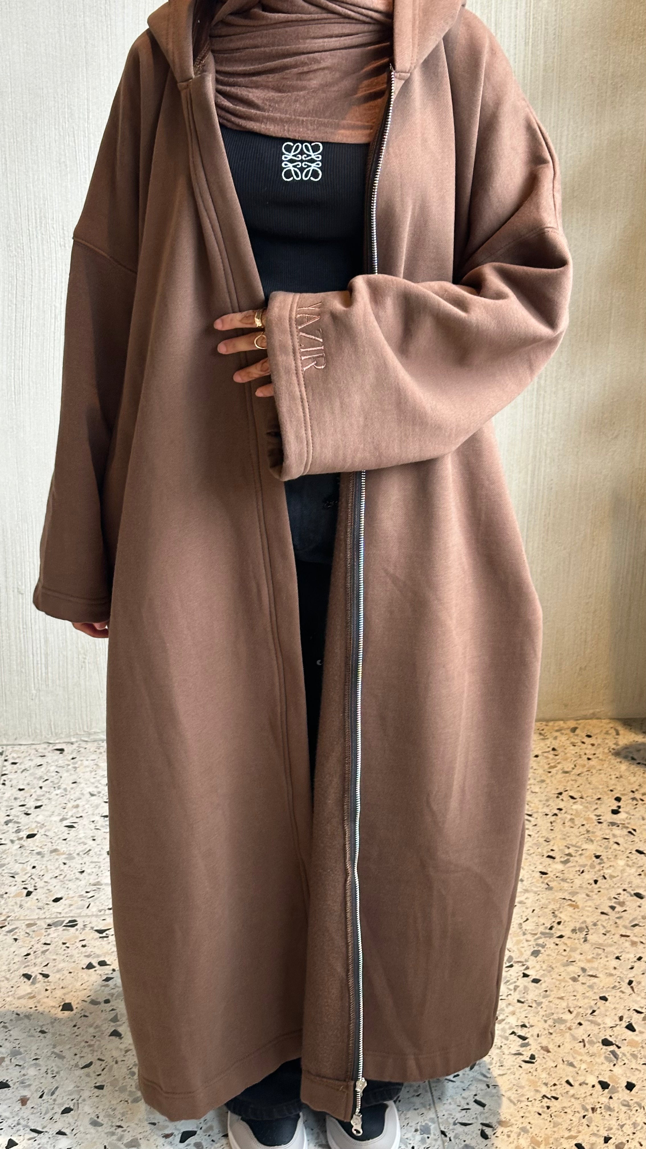 A person wearing a long, loose-fitting taupe-brown hooded zip-front abaya or long cardigan over a black top and matching hijab, standing indoors on a speckled terrazzo floor against a light textured wall.