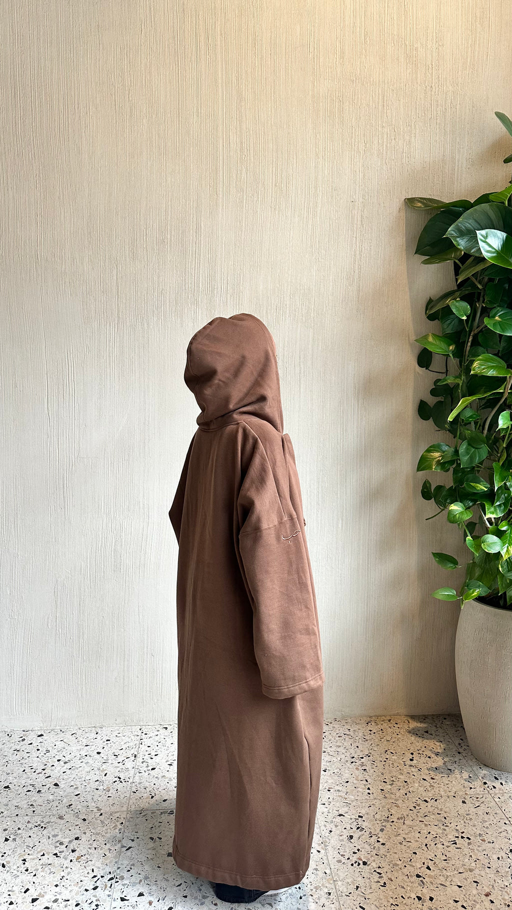 A person wearing a long, loose-fitting brown hooded abaya robe stands in profile against a textured beige wall, with a green potted plant on the right and a speckled floor below.