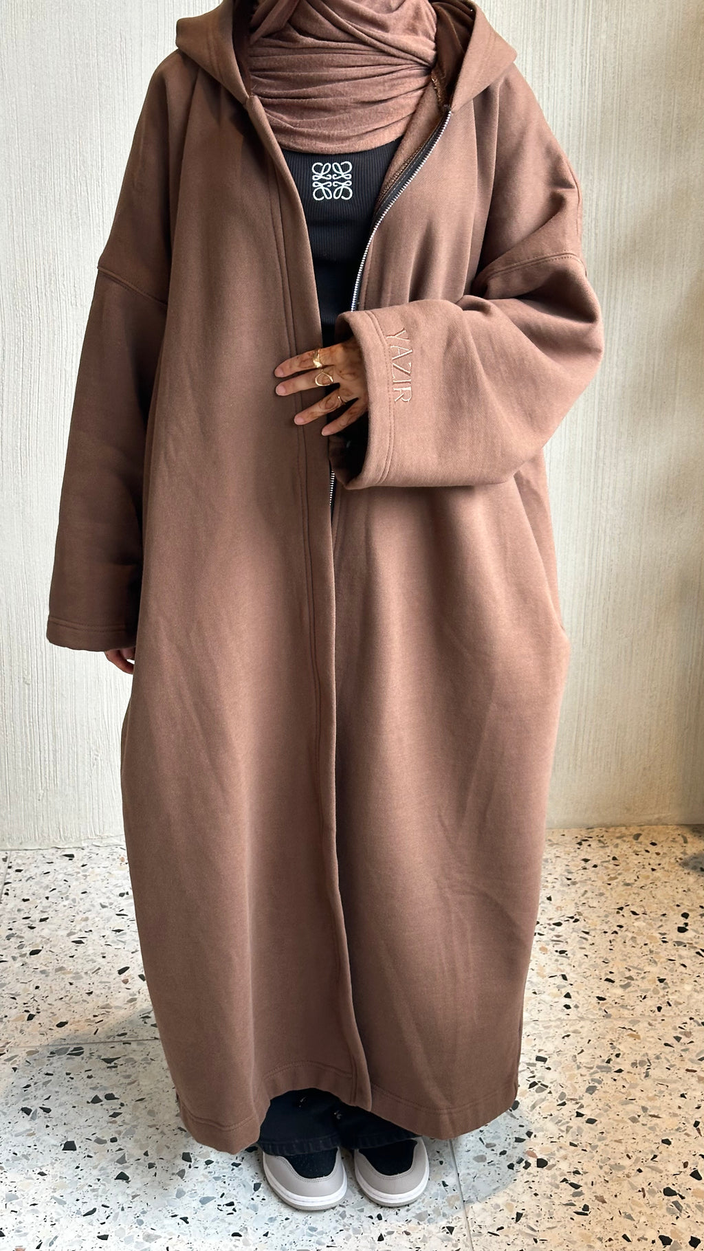 A person wearing a long, oversized taupe-brown hooded coat over a black top and matching hijab, standing indoors on a speckled floor against a light textured wall. Perfect for winter abaya