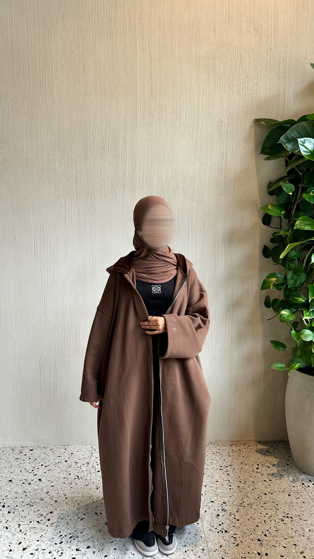 A person wearing a long taupe-brown abaya with a front zipper, a matching brown hijab, and a black top, standing against a textured beige wall with a potted green plant on the right.
