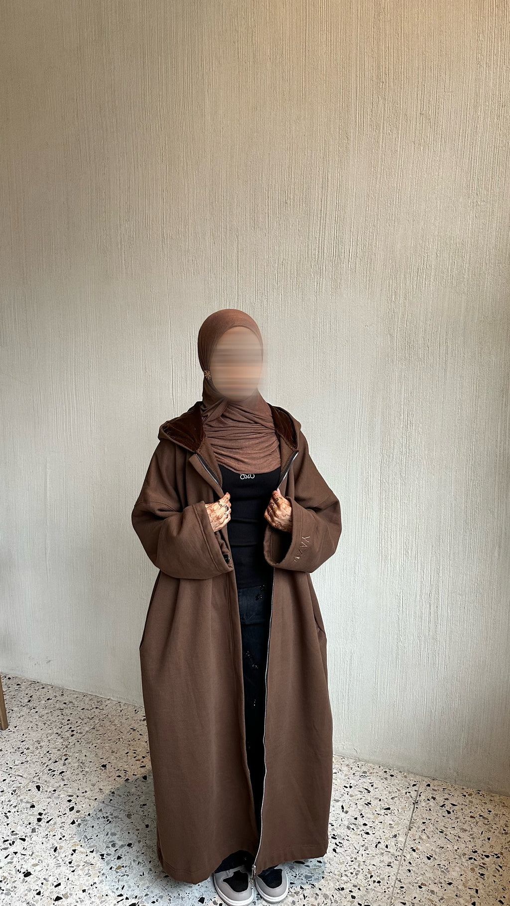 A person wearing a long brown hooded coat over a black outfit and brown hijab, standing against a textured beige wall with a speckled floor.
