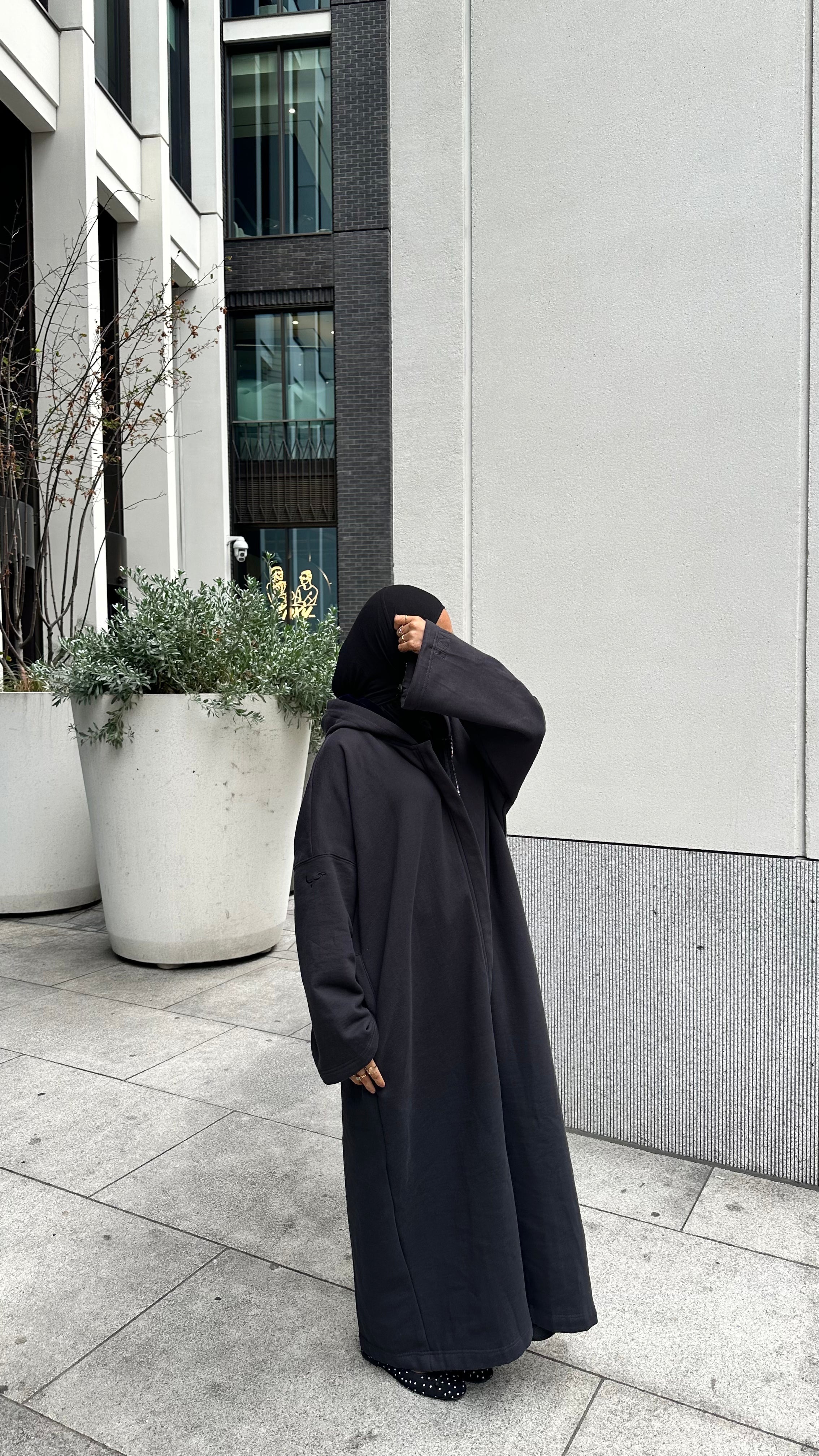 A person wearing a long black hooded abaya and a black hijab stands on a paved outdoor walkway in front of a modern building with large white planters. Black winter abaya 