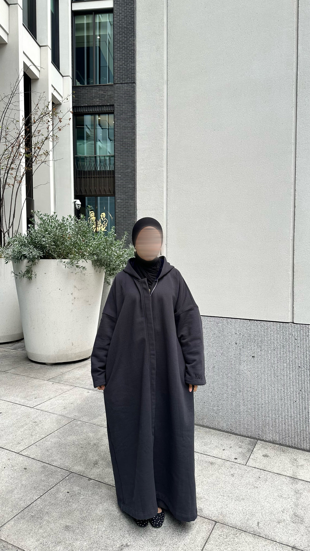 A person wearing a long, loose-fitting black abaya with a hood and black shoes, standing outdoors on a paved walkway in front of a modern building. Winter abaya black with matching hijab